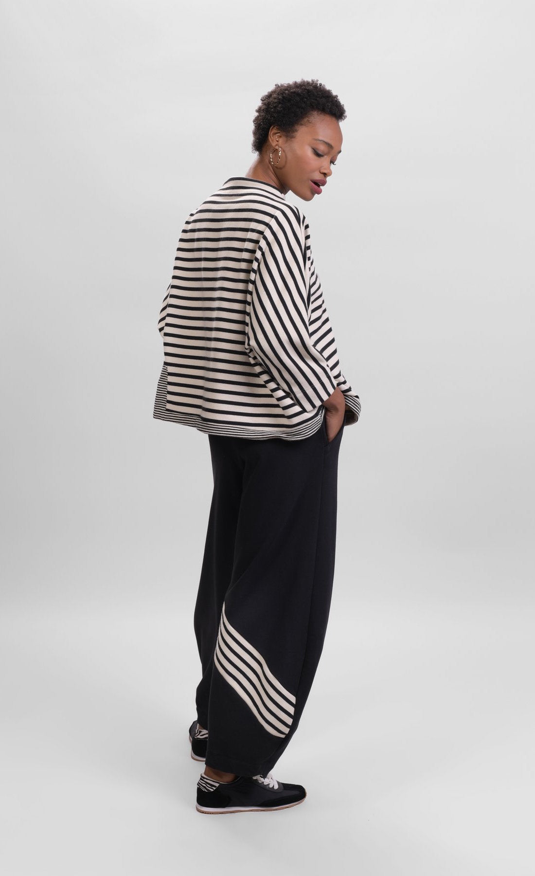 Back full body view of a woman wearing the Alembika Urban French Terry Pants with the Alembika Urban French Terry Stripes Top. This top is white with black striping all over it. The top has an asymmetrical hem, long sleeves, a mock neck, and a boxy silhouette.