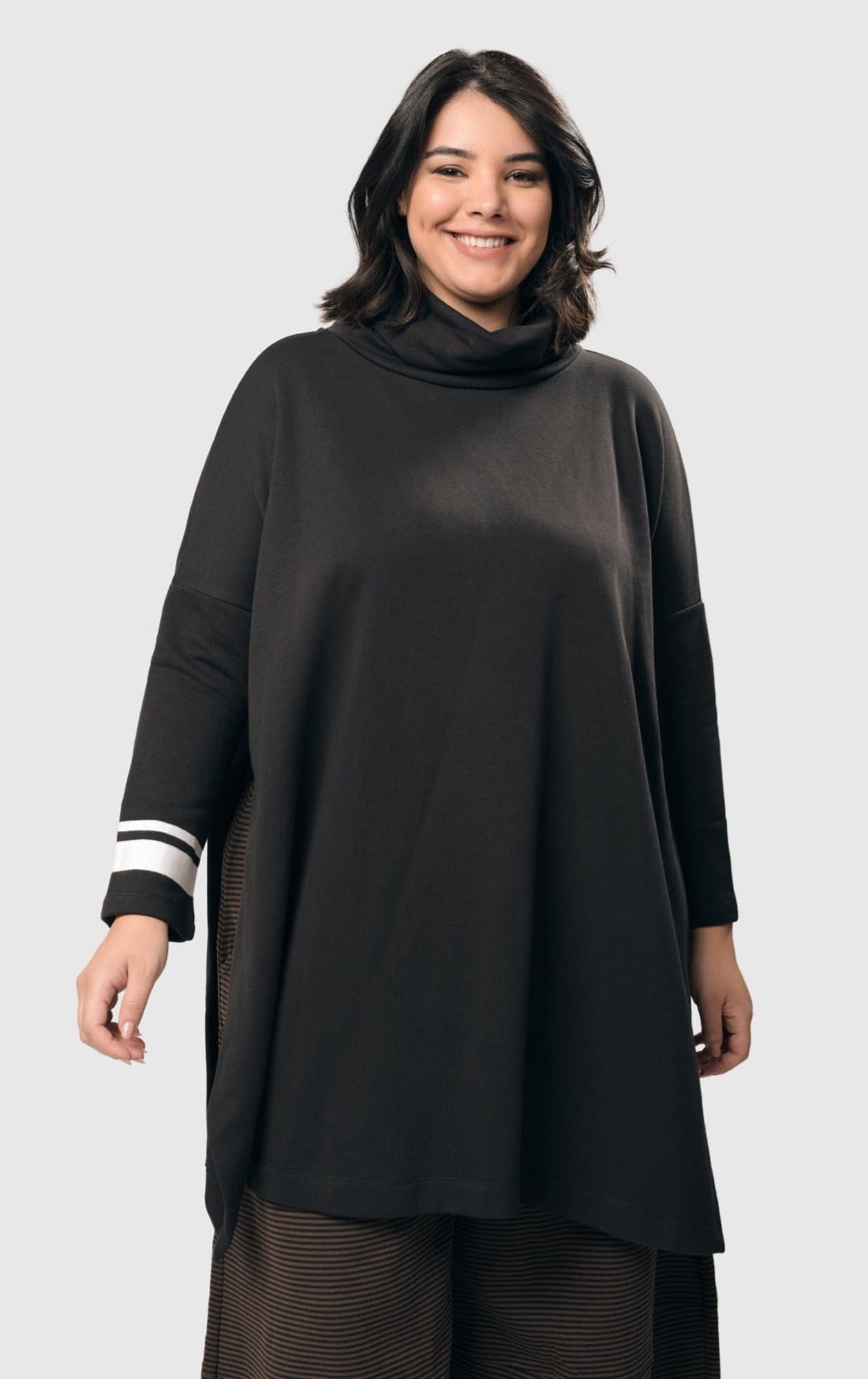 Front view of a woman wearing the alembika urban ribbon cowl neck tunic top in black. This tunic has deep side slits, a cowl neck, and long sleeves with two white stripes on the right arm.