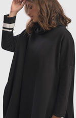 Load image into Gallery viewer, Front view of a woman wearing the alembika urban ribbon cowl neck tunic top in black. This tunic has deep side slits, a cowl neck, and long sleeves with two white stripes on the right arm.
