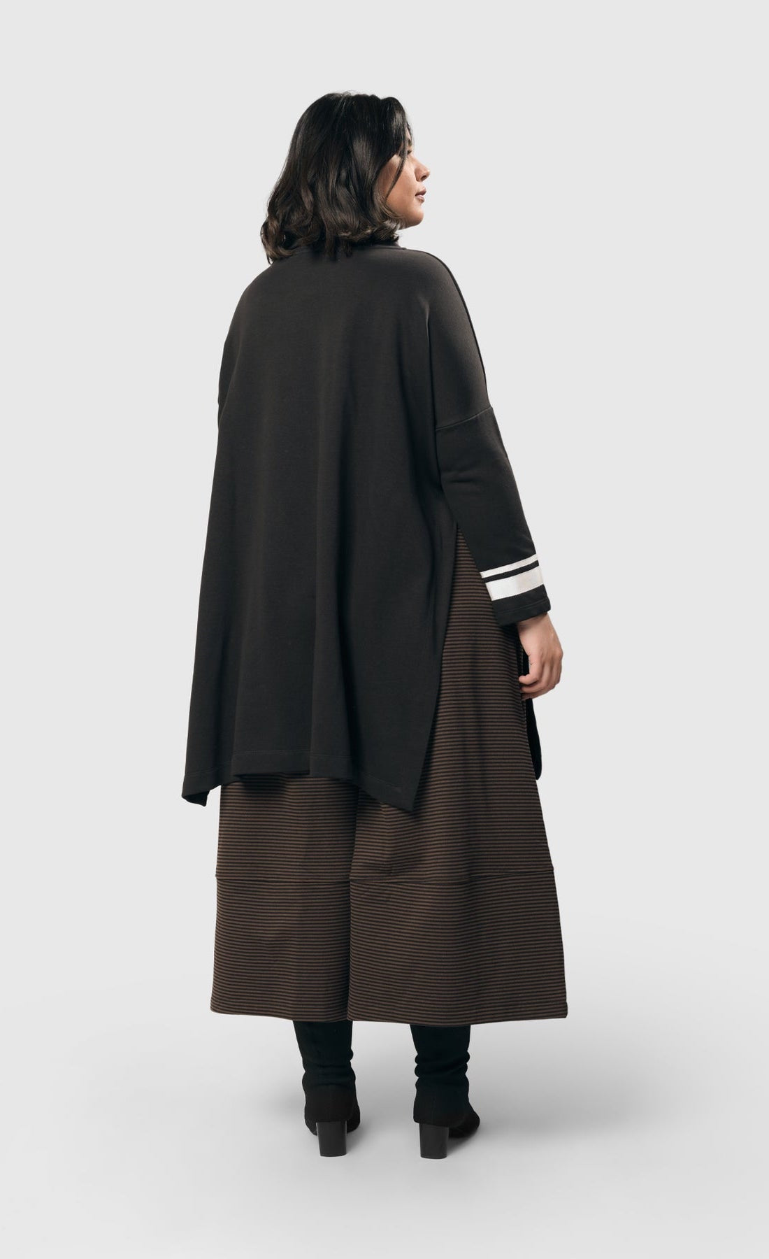 Back view of a woman wearing the alembika urban ribbon cowl neck tunic top in black. This tunic has deep side slits, a cowl neck, and long sleeves with two white stripes on the right arm.