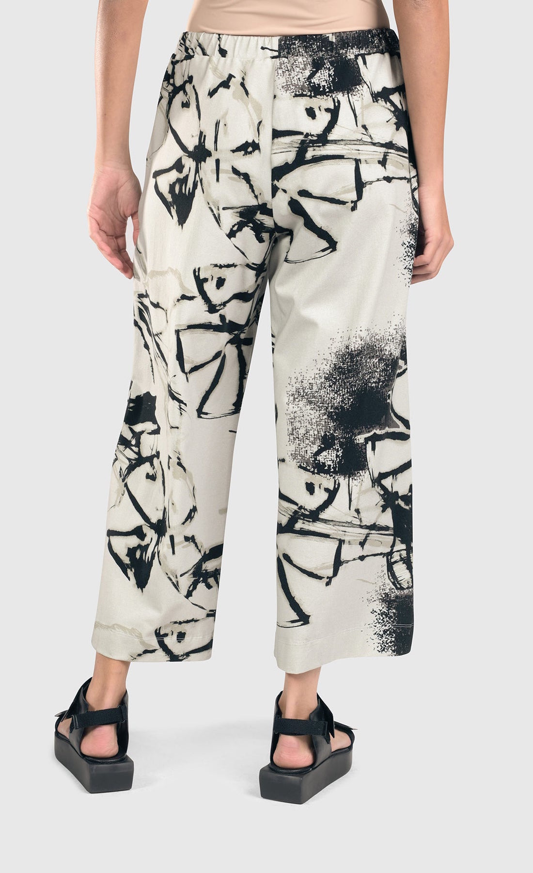 Back bottom half view of a woman wearing the alembika urban sketch go to pant. This pant is grey white with black and dark grey scribbles all over it. The pant has a cropped cut, and wide legs.