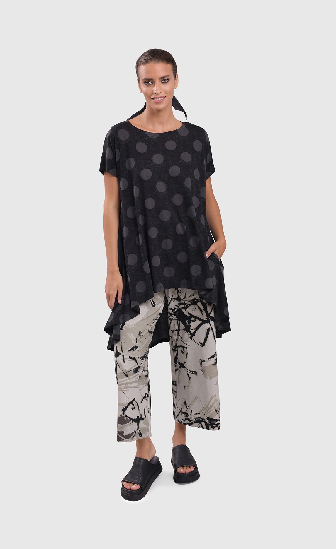 Front full body view of a woman wearing an alembika dotted top and the alembika urban sketch go to pant. This pant is grey white with black and dark grey scribbles all over it. The pant has a cropped cut, and wide legs.