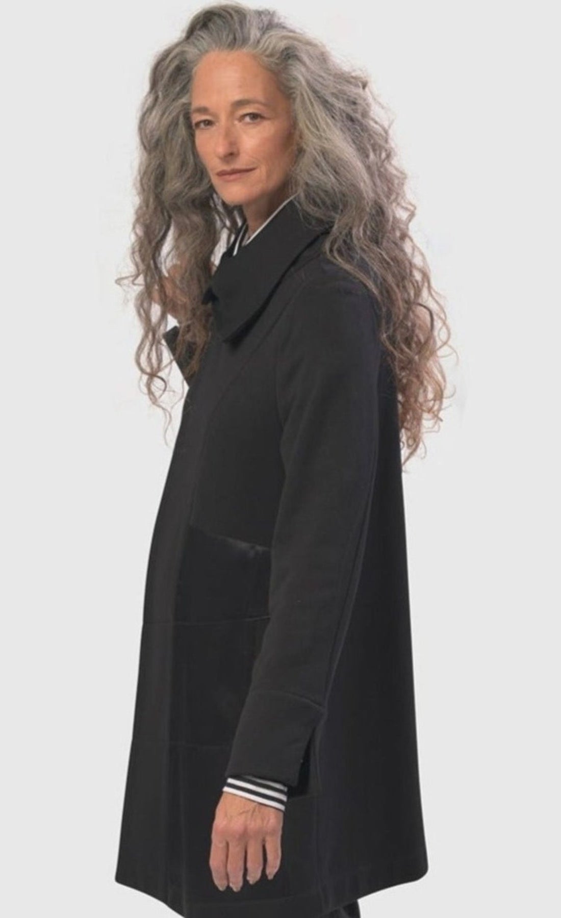 Left side top half view of a woman wearing the alembika urban sleek satin trim jacket in black. This jacket has a single button closure and satin panels on the side. 