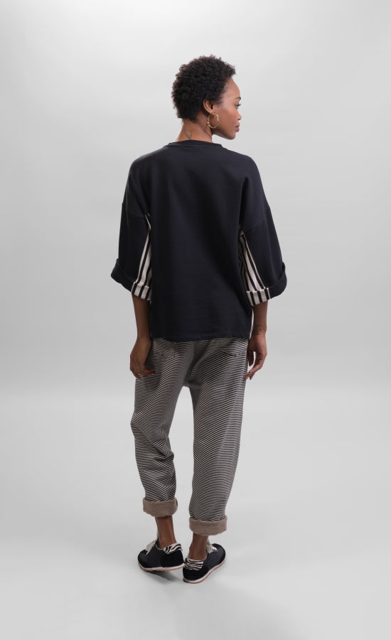 Back full body view of a woman wearing striped pants and the alembika urban black/striped panel top. This top is black with wide folded sleeves that sit at the elbows. The top has black and white striped paneling on the sides and under the arms.