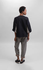Load image into Gallery viewer, Back full body view of a woman wearing striped pants and the alembika urban black/striped panel top. This top is black with wide folded sleeves that sit at the elbows. The top has black and white striped paneling on the sides and under the arms.

