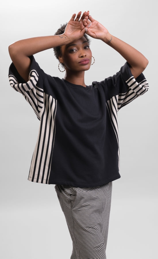 Front top half view of a woman with her hands resting on her forehead and wearing striped pants and the alembika urban black/striped panel top. This top is black with wide folded sleeves that sit at the elbows. The top has black and white striped paneling on the sides and under the arms.