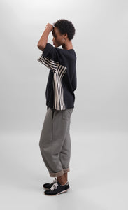 Left side full body view of a woman wearing striped pants and the alembika urban black/striped panel top. This top is black with wide folded sleeves that sit at the elbows. The top has black and white striped paneling on the sides and under the arms.