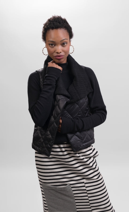 Front top half view of a woman wearing a black and white striped skirt and the alembika urban puffer vest in black. This vest has a ribbed collar and a front single button.