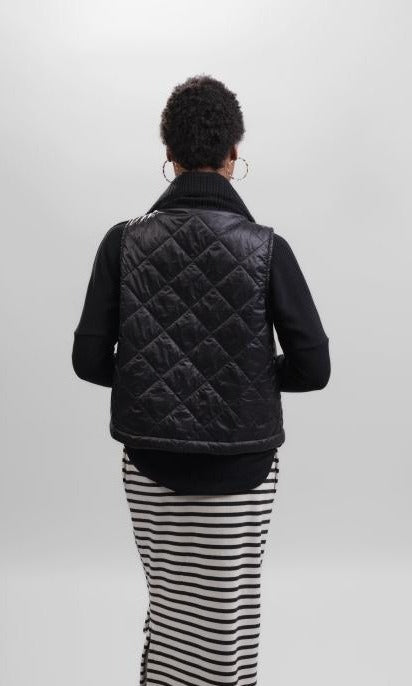 Back top half view of a woman wearing a black and white striped skirt and the alembika urban puffer vest in black. This vest has a ribbed collar.