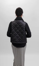 Load image into Gallery viewer, Back top half view of a woman wearing a black and white striped skirt and the alembika urban puffer vest in black. This vest has a ribbed collar.
