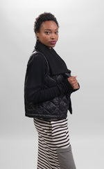 Load image into Gallery viewer, Right side top half view of a woman wearing a black and white striped skirt and the alembika urban puffer vest in black over a black long sleeve. This vest has a ribbed collar and a front single button.
