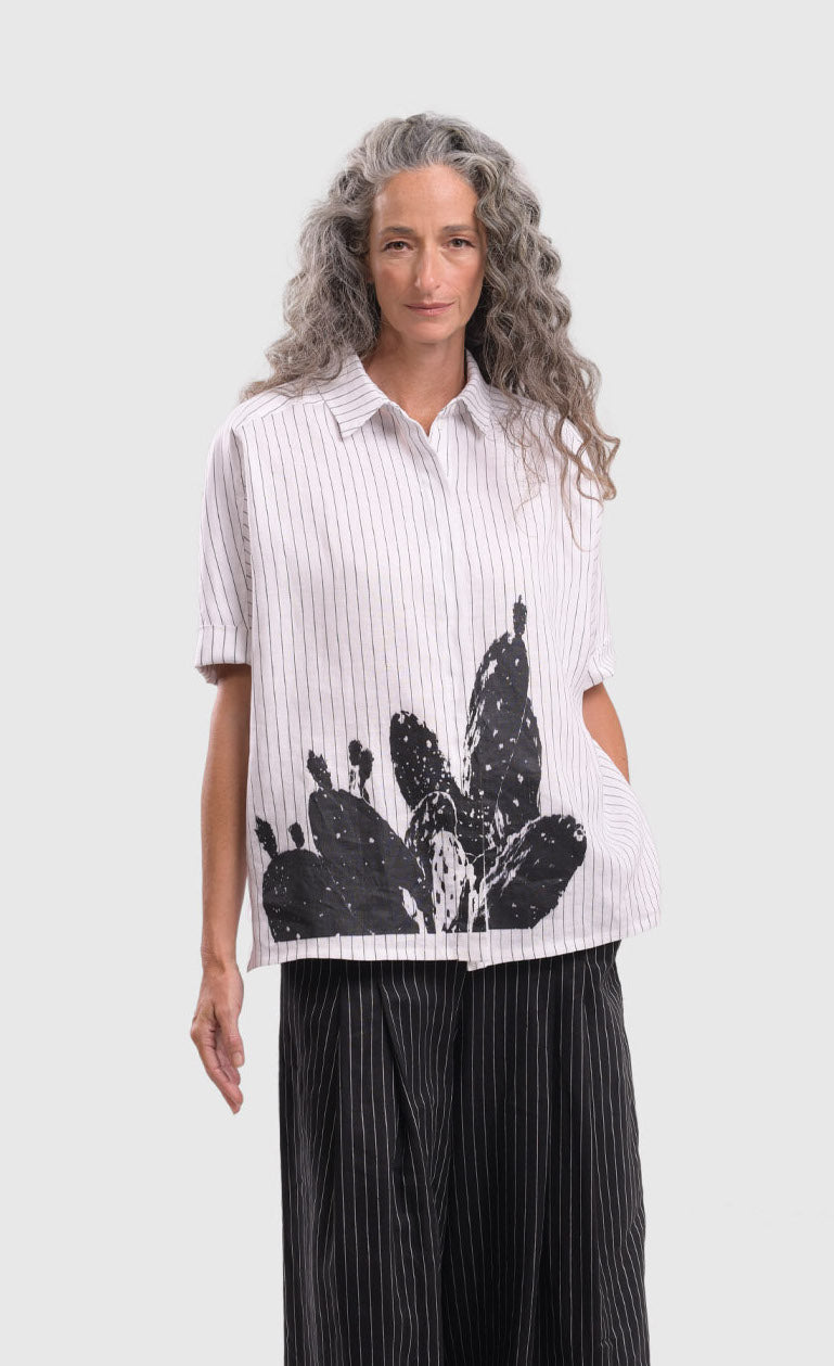 Front top half view of a woman wearing the alembika striped white cactus shirt. This white shirt has black pinstripes, a button down front, short sleeves, a collar, and a black cactus print on the bottom of the front.