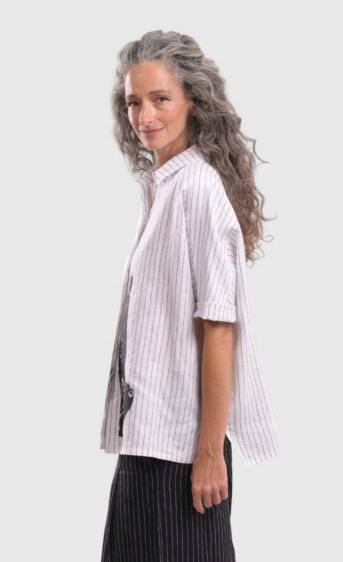 Left side top half view of a woman wearing the alembika striped white cactus shirt. This white shirt has black pinstripes, a button down front, short sleeves, a collar, and a black cactus print on the bottom of the front.