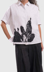 Close up front view of a woman wearing the alembika striped white cactus shirt. This white shirt has black pinstripes, a button down front, short sleeves, a collar, and a black cactus print on the bottom of the front.