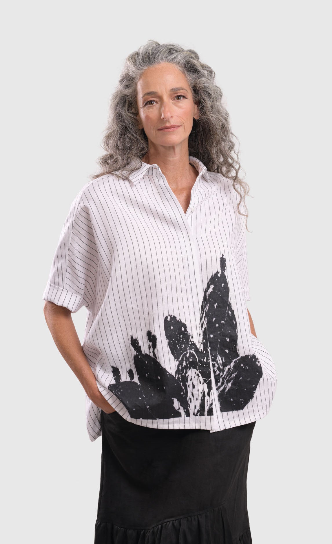 Front top half view of a woman wearing the alembika striped white cactus shirt. This white shirt has black pinstripes, a button down front, short sleeves, a collar, and a black cactus print on the bottom of the front.