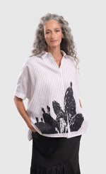 Load image into Gallery viewer, Front top half view of a woman wearing the alembika striped white cactus shirt. This white shirt has black pinstripes, a button down front, short sleeves, a collar, and a black cactus print on the bottom of the front.
