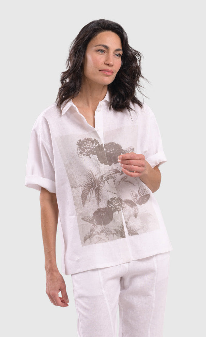 Front top half view of a woman wearing the alembika white flowers shirt. This shirt is white with a screen print of monochrome flowers on a square on the front. The shirt has short sleeves that are rolled up, a button down front, and a shirt collar. 