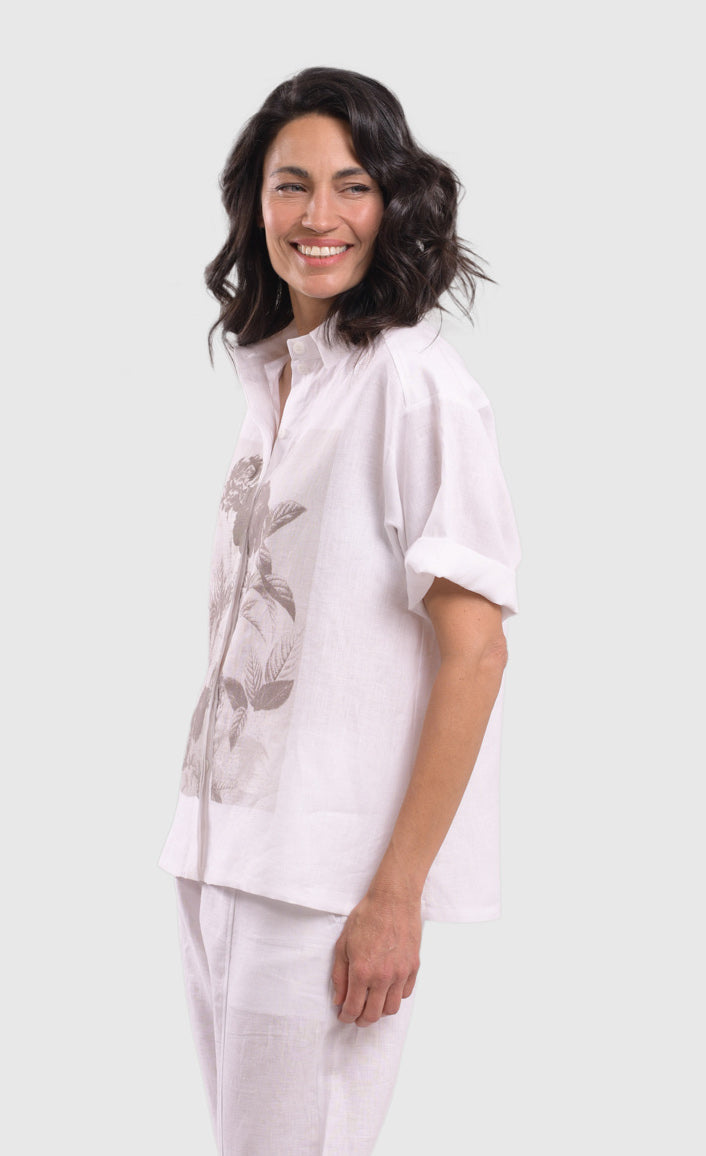 Left side top half view of a woman wearing the alembika white flowers shirt. This shirt is white with a screen print of monochrome flowers on a square on the front. The shirt has short sleeves that are rolled up, a button down front, and a shirt collar. 