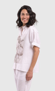Left side top half view of a woman wearing the alembika white flowers shirt. This shirt is white with a screen print of monochrome flowers on a square on the front. The shirt has short sleeves that are rolled up, a button down front, and a shirt collar. 