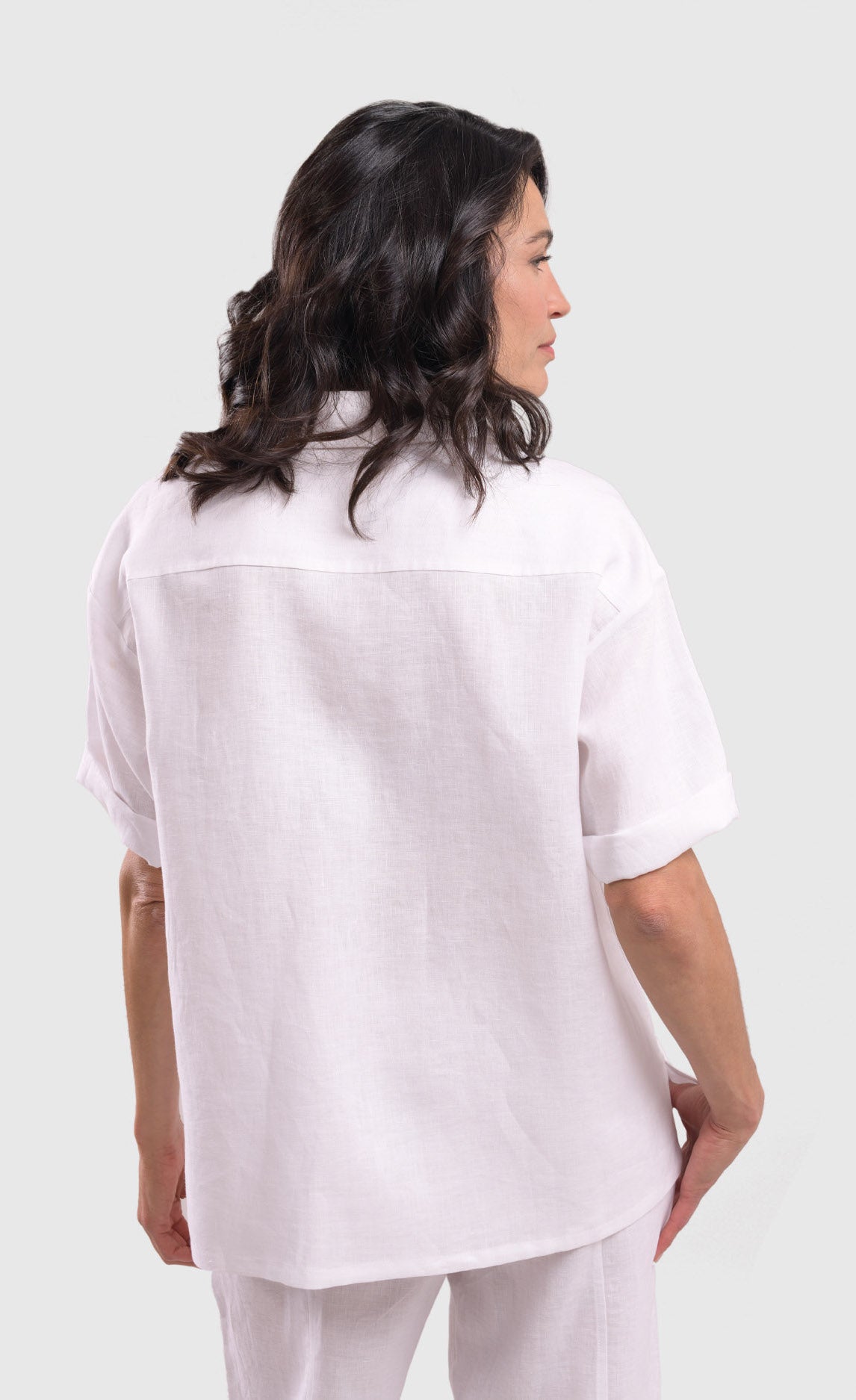 Back top half view of a woman wearing the alembika white flowers shirt. This shirt is white with a screen print of monochrome flowers on a square on the front. The shirt has short sleeves that are rolled up, a button down front, and a shirt collar. 