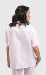 Load image into Gallery viewer, Back top half view of a woman wearing the alembika white flowers shirt. This shirt is white with a screen print of monochrome flowers on a square on the front. The shirt has short sleeves that are rolled up, a button down front, and a shirt collar. 
