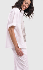 Load image into Gallery viewer, Right side top half view of a woman wearing the alembika white flowers shirt. This shirt is white with a screen print of monochrome flowers on a square on the front. The shirt has short sleeves that are rolled up, a button down front, and a shirt collar. 
