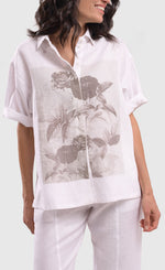 Load image into Gallery viewer, Front close up view of a woman wearing the alembika white flowers shirt. This shirt is white with a screen print of monochrome flowers on a square on the front. The shirt has short sleeves that are rolled up, a button down front, and a shirt collar. 
