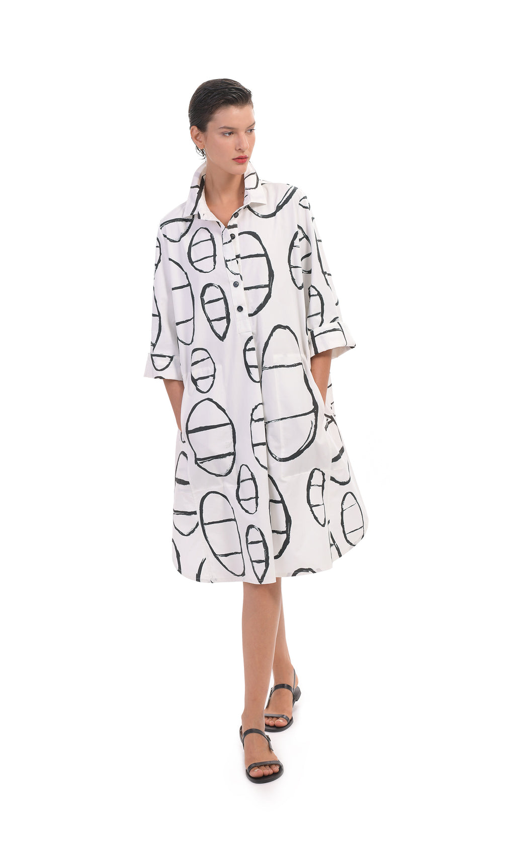 Front full body view of a woman wearing the alembika cotton print shirt. This shirt goes down to the knees and has elbow length sleeves. It features a button up front with a collar. It is white with a black circle/capsule like print all over it.