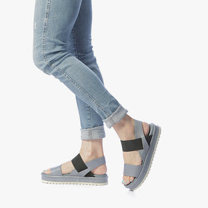 outer and inner view of a woman wearing a pair of the all black banded trekker. This sandal has a strap over the toe bed and behind the heel with a black elastic strap over the instep. The rest of the shoe is a blue leather. 