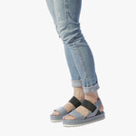 Load image into Gallery viewer, outer view of a woman wearing a pair of the all black banded trekker. This sandal has a strap over the toe bed and behind the heel with a black elastic strap over the instep. The rest of the shoe is a blue leather.
