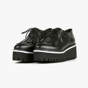 Front view of a pair of the all black footwear kiltie ox flatform. This shoe is black with a black platform sole and a lace up front. The sole and leather upper are separated by white piping around the shoe.
