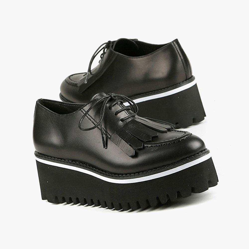 Outer and inner side view of a pair of the all black footwear kiltie ox flatform. This shoe is black with a black platform sole and a lace up front. The sole and leather upper are separated by white piping around the shoe.