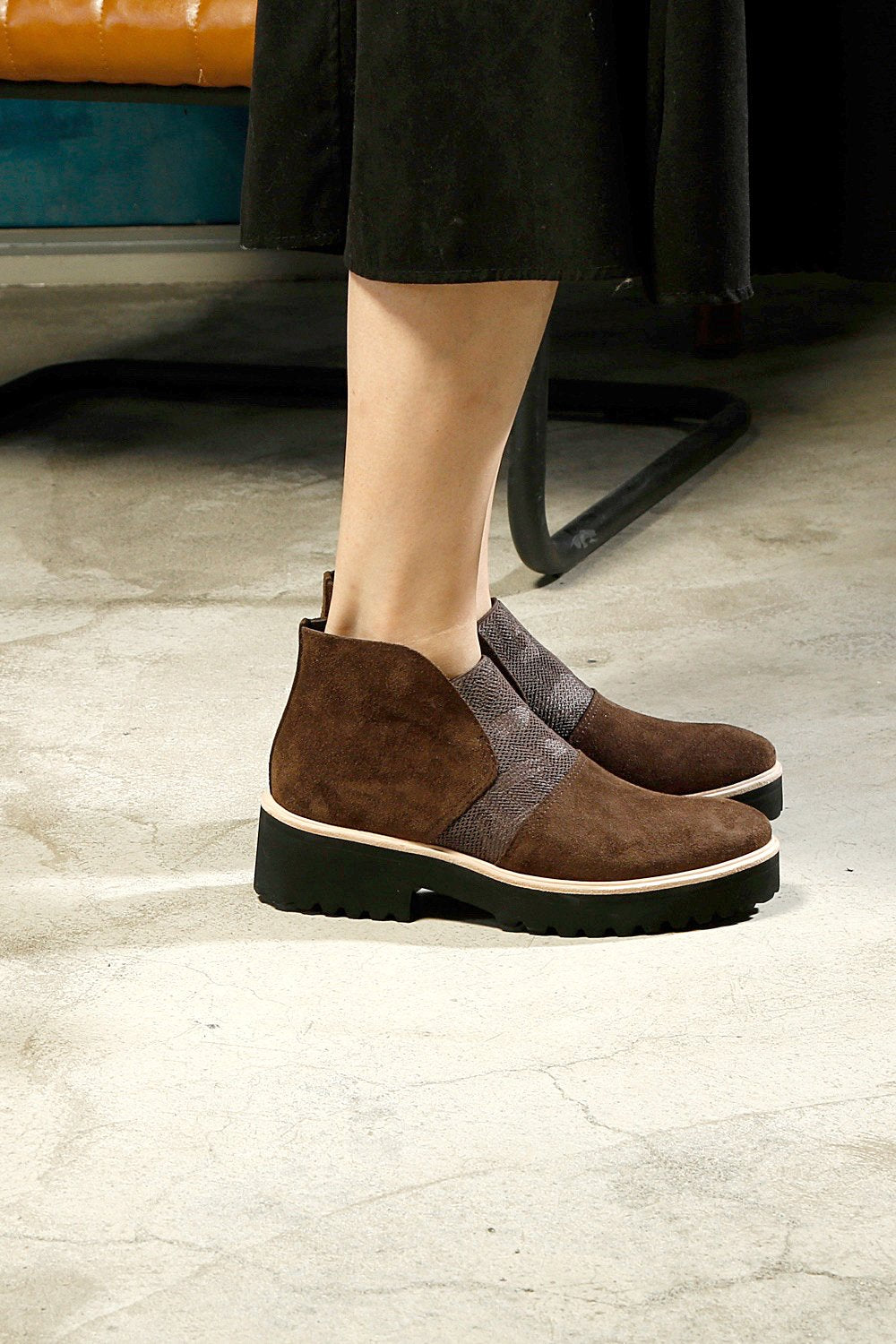 Outer view of a woman wearing a pair of the all black nu banded bootie in brown. This ankle length bootie has a suede upper with an elastic camouflage inlet. The lugg sole is black with white piping.