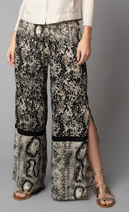 Front, bottom half view of a woman wearing the Lola & Sophie Animal Border Print Pull-On Pant. These pants feature a smudge print and a snake print separated by a back stripe that runs horizontally across the leg near the knee. These relaxed, straight pants have a side slit that goes up to the knee.
