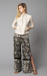 Load image into Gallery viewer, Front, full body view of a woman with her hands in the front pockets of the beige button up jacket she is wearing. On the bottom-half, she is wearing the Lola &amp; Sophie Animal Border Print Pull-On Pant. These pants feature a smudge print and a snake print separated by a back stripe that runs horizontally across the leg near the knee. These relaxed, straight pants have a side slit that goes up to the knee.
