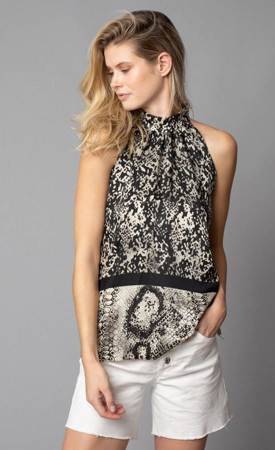 Front, top half view of a woman with her left hand in the left pocket of her white shorts. On the top, the woman is wearing the lola & sophie border animal print halter top. This sleeveless top has a high halter neck and a mix of two different snake skin prints separated by a black border that runs horizontally across the waist.