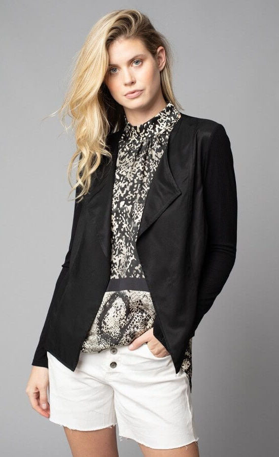 Front, top half view of a woman with her left hand in the left pocket of her white shorts. On the top, the woman is wearing the lola & sophie black Crinkle Linen Zip-Front Long Jacket over the lola & sophie border animal print halter top. The tank underneath has a high halter neck and a mix of two different snake skin prints separated by a black border that runs horizontally across the waist.