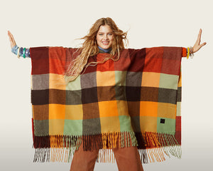 Front top half view of a woman wearing the desigual plaid poncho with brown pants. This poncho is brown, red, orange, and green plaid. It has a hood and a fringed hem.