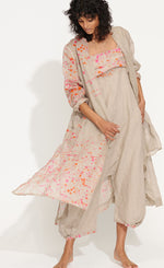Load image into Gallery viewer, front full body view of a woman wearing the banana blue bright flax splash print duster over a matching jumpsuit. This duster is flax/beige colored with pink and orange splatter paint print. This duster goes down to the knees, has long sleeves, a straight fit, and a short stand collar.
