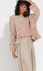 Load image into Gallery viewer, Front top half   view of a woman wearing the banana blue bright flax splash top. This top is flax colored with a pink and orange splatter painted print appearance and a boxy silhouette. This top also has long sleeves and a boat neck.
