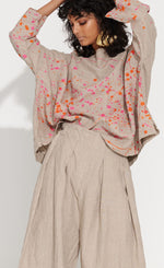 Load image into Gallery viewer, Front top half   view of a woman wearing the banana blue bright flax splash top. This top is flax colored with a pink and orange splatter painted print appearance and a boxy silhouette. This top also has long sleeves and a boat neck.

