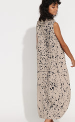 Load image into Gallery viewer, Back full body view of a woman wearing the banana blue navy splash flax dress. This dress is flax/beige colored with a navy splatter paint print all over it. The dress is sleeveless with a cowl neck and a balloon like shape.
