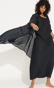 Front full body view of a woman wearing the banana blue solid navy splash duster. This duster is a dark navy that looks black. Under the duster the model is wearing a matching top and bottom. The duster has long sleeves, layered collar, and a pleated seam detail.