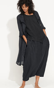 Front full body view of a woman wearing the banana blue solid navy splash duster. This duster is a dark navy that looks black. Under the duster the model is wearing a matching top and bottom. The duster has long sleeves, layered collar, and a pleated seam detail.