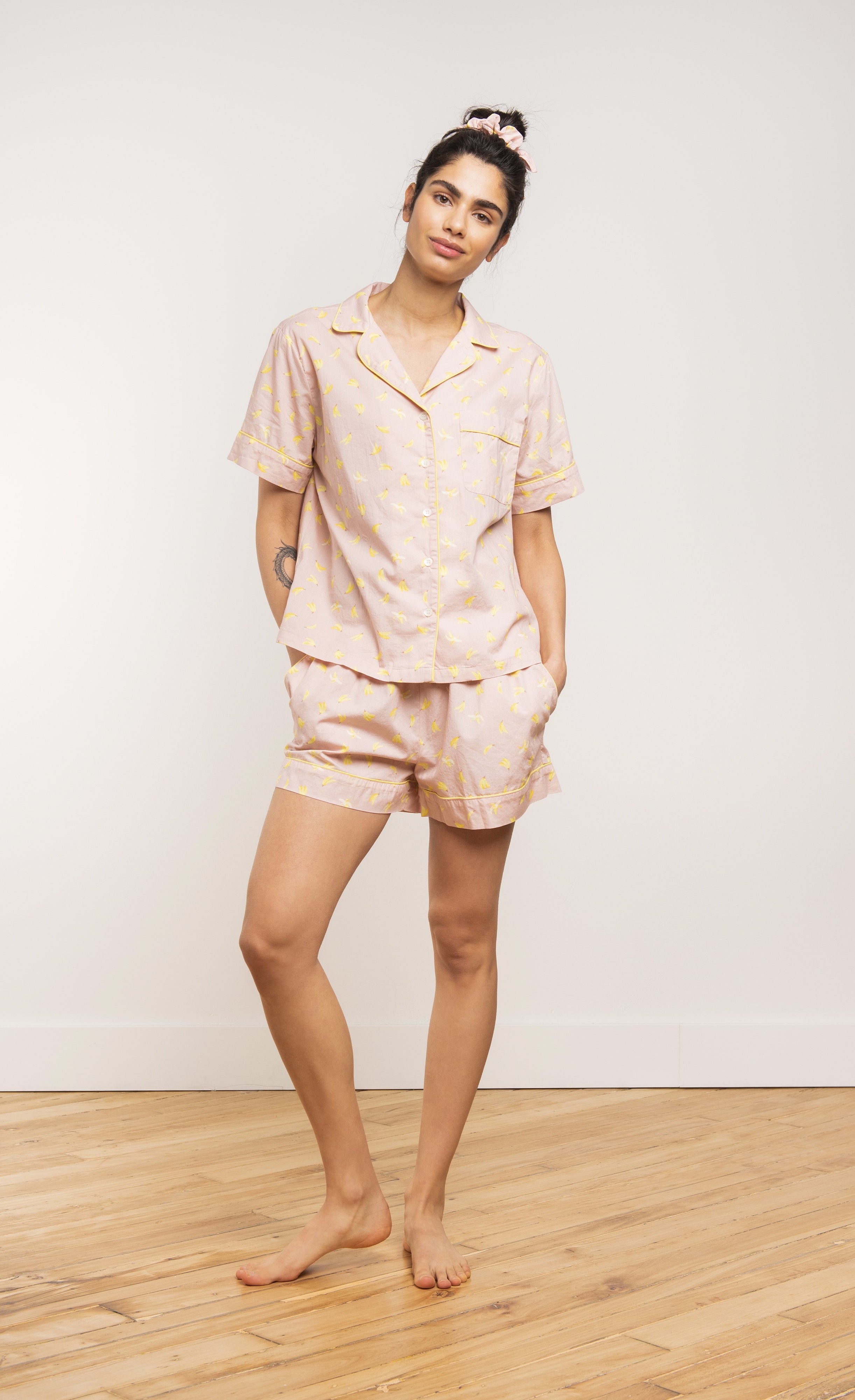 Front view of a woman wearing the coffee shoppe banana pj set. This set is pink with a banana print all over it. . The top is a button down short sleeve and the bottom are shorts.