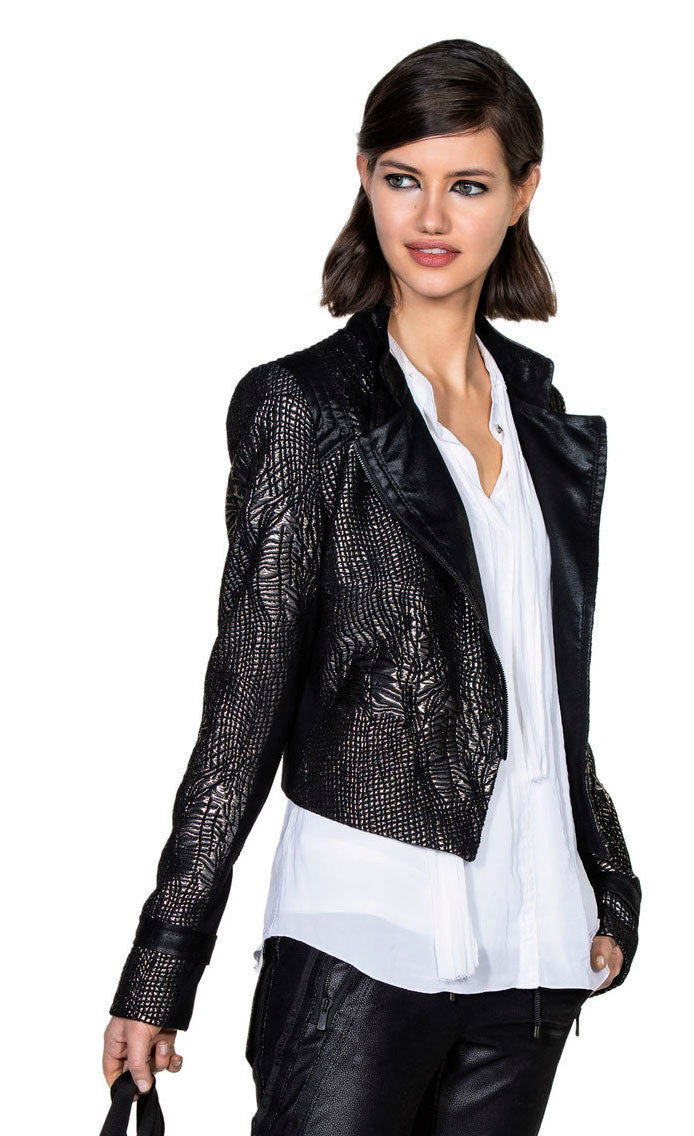 Front top half view of a woman wearing the beate heymann carbon jacket. This jacket is black with a cracked gold print all over it. The jacket is short with long sleeves, and off-center zipper, and a notched collar.