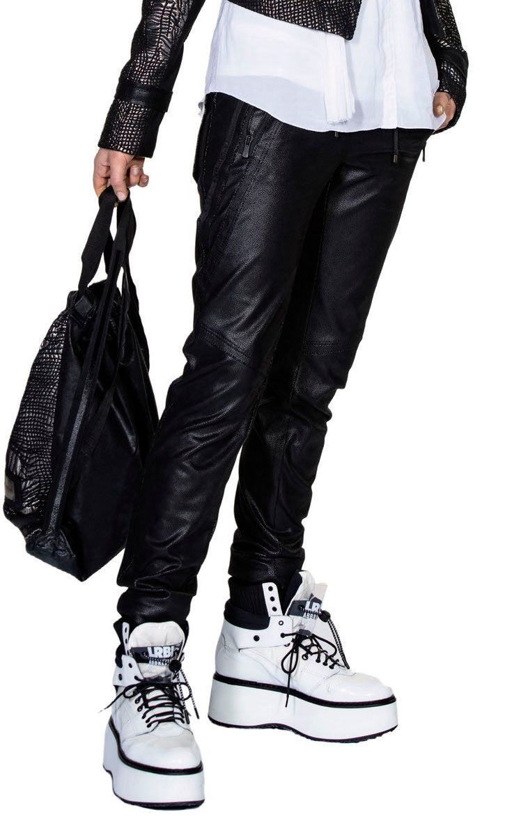 Front bottom half view of a model wearing the beate heymann signature jogger. These joggers are black and appear faux leather. They have decorative stitching all over the leg and two front zip pockets.