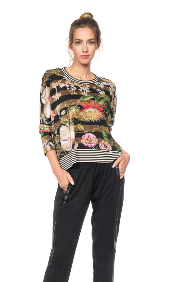 Front top half view of a woman wearing black joggers and the Beate Heymann Floral Striped Top. This top is striped black and gold with flowers on top of it. The neck and the hem have black and white striped trim. The top also has long sleeves.