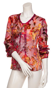 Front top half view of a mannequin wearing the beate heymann fuchsia firewall blouse. This blouse has a mixed red, pink, and orange print and is see through. The top also has long sleeves, a front inverted pleat, and a round neck.