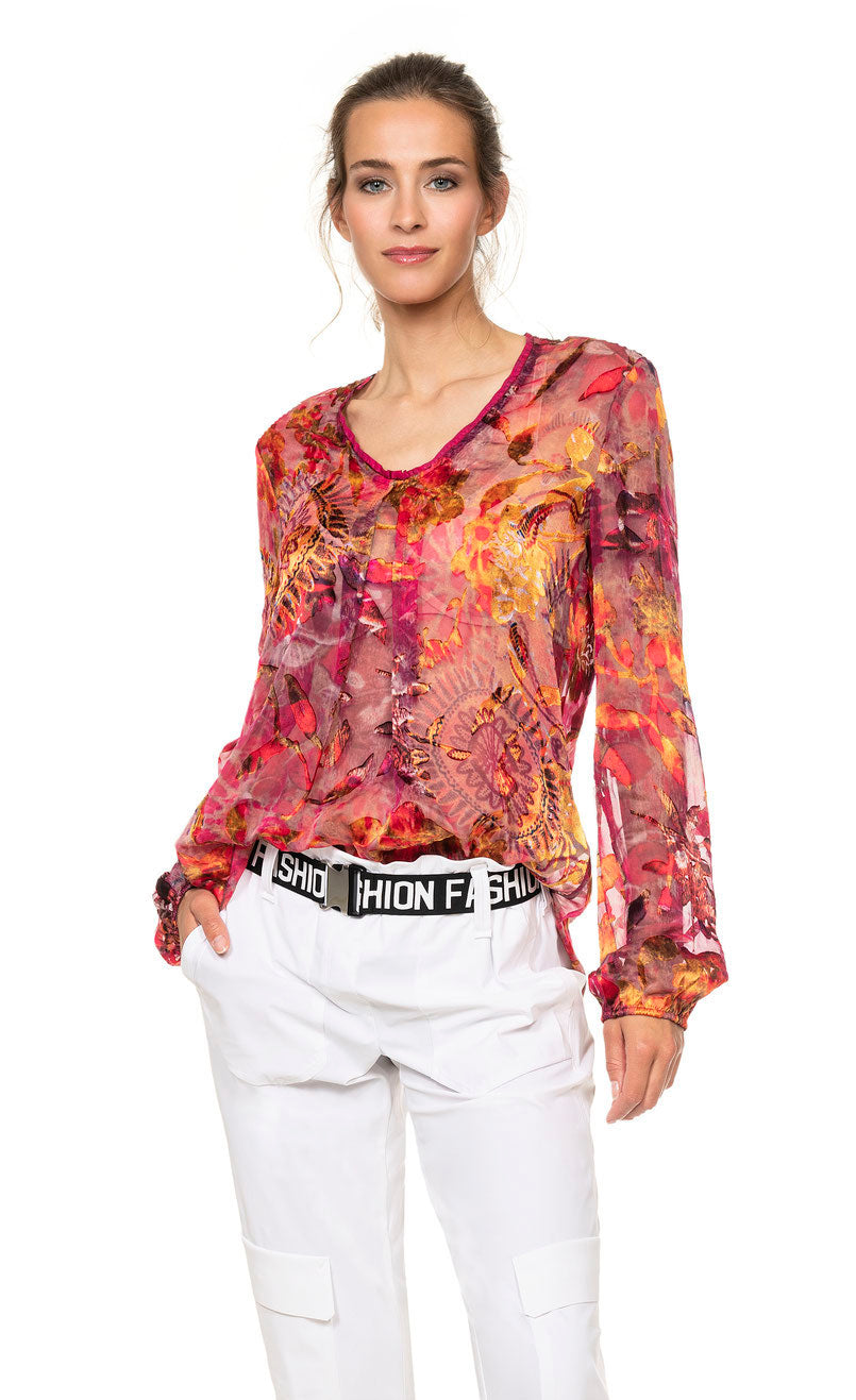 Front top half view of a woman wearing the beate heymann fuchsia firewall blouse. This blouse has a mixed red, pink, and orange print and is see through. The top also has long sleeves, a front inverted pleat, and a round neck.